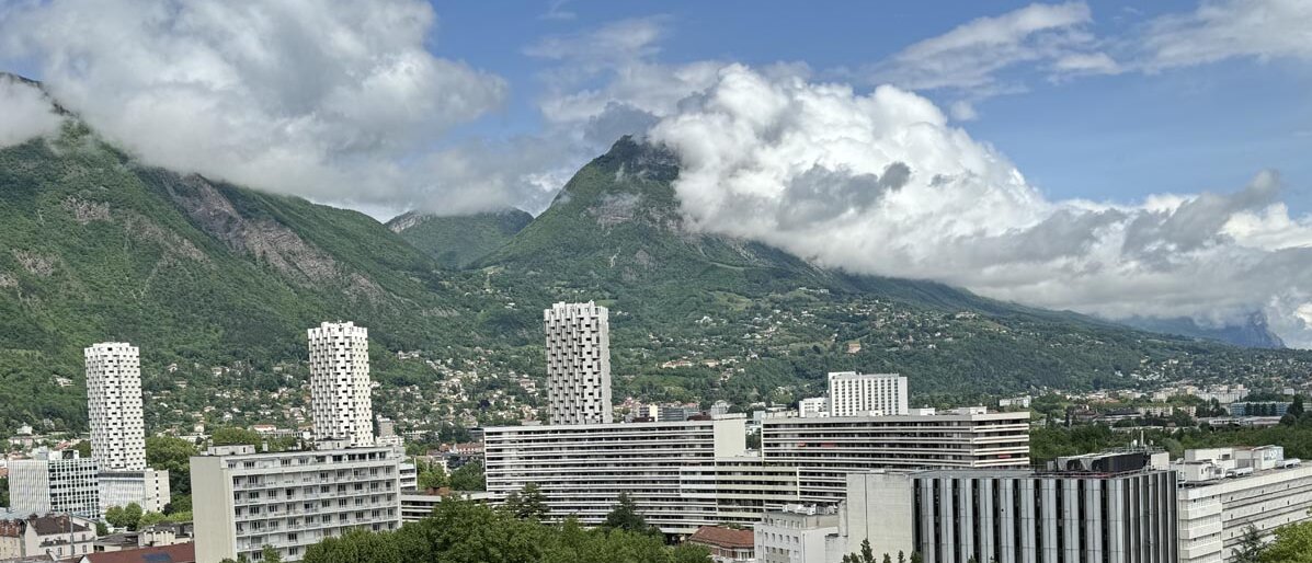 Drei Hochhäuser, mehrere Wohnblöcke, wolkenbehangenes Gebirge im Hintergrund, befahrene Straße, viele Häuser auf dem Hang des Berges