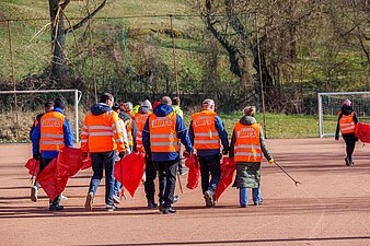 Auf dem Foto sieht man mehrere Mitarbeiter von hinten mit Greifzangen und Müllsäcken in der Hand laufen. Sie haben Warnwesten mit der Aufschrift „Mit uns läuft´s“ an.