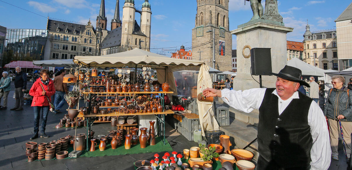 Töpfer handwerker mit schwarzer Weste mit einladender Geste und Töpferstand im Hintergrund