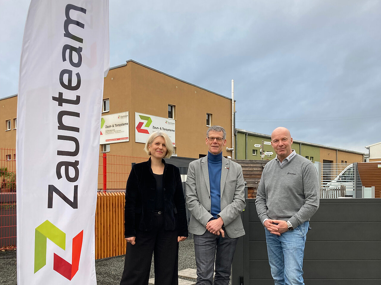 Eine Frau und zwei Männer stehen neben einer Fahne vor einem Firmengebäude