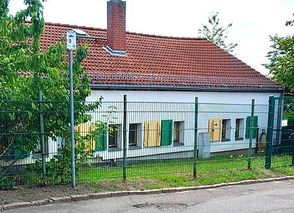 helles eingeschossiges Gebäude mit rotem Dach und bunten Fensterläden, davor eine kleine Grünfläche, zum Fußweg abgegrenzt durch einen Metallzaun, links steht ein Laubbaum