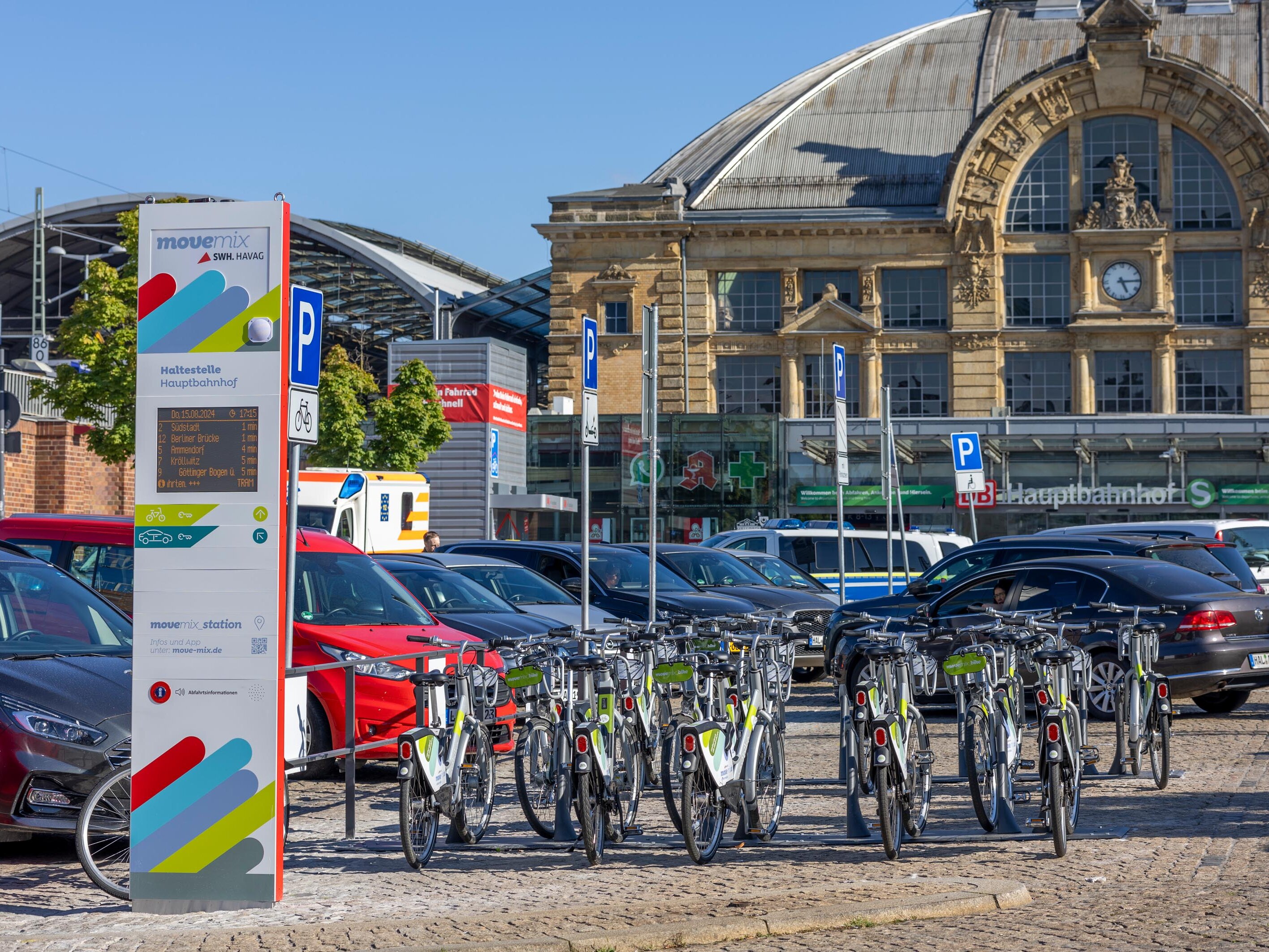 Das Bild zeigt den Hauptbahnhof Halle (Saale) im Hintergrund. Im Vordergrund befinden sich abgestellte Fahrräder, eine elektronische Anzeigetafel der HAVAG und parkende Autos.