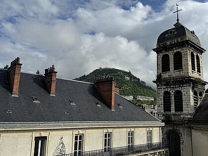 Haus mit vielen Fenstern, Turm, Berg im Hintergrund mit Bastille drauf