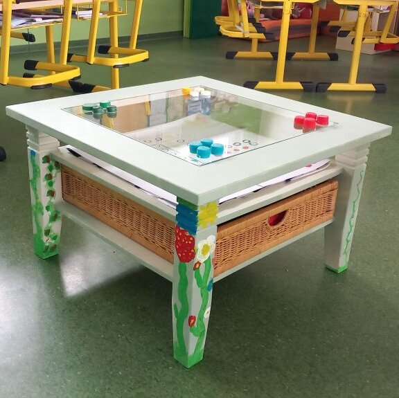 Ein Holztisch wurde mit Blumen bemalt. Große weiße Blätter mit Zeichnungen sind die Spiele. Flaschendeckel sind die Spieler.