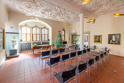 Weiß gestrichener Raum mit reich verzierter Stuckdecke und roten Bodenfliesen. Im Hintergrund drei nebeneinander liegende Fenster mit Butzenscheiben. Im Raum sind drei Stuhlreihen mir grauen Stühlen aufgebaut. Davor ein Tisch mit zwei Stühlen für das Brautpaar.