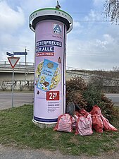 An einer Litfaßsäule stehen befüllte Müllsäcke sowie ausgediente Weihnachtsbäume. 