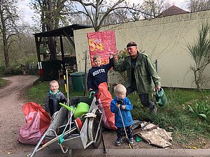 Ein Mann mit drei Kindern, die als Gruppe mit einem Anhängern, Müllsäcken und Geräten vor einem gebastelten Aktionsaufruf stehen.