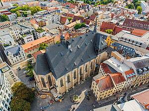 Sandsteinfarbene Kirche mit dunklem Dach von oben aus der Luft fotografiert. Vor der Kirche ein kleiner Springbrunnen. Um die Kirche städtische Bebauung mit Wohn- und Geschäftshäusern.