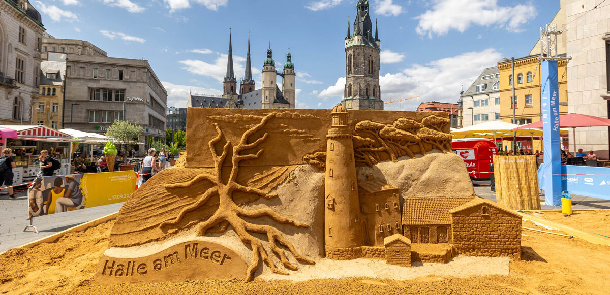 Eine aus Sand gebaute Skulptur auf dem Marktplatz mit Leuchtturm, Gebäude und Bäumen, im Hintergrund die 5 Türme von Halle