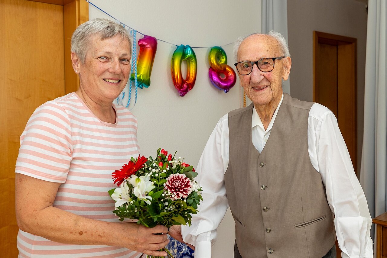 Beide stehen in einer Wohnungm Fr. Brederlow hält einen Blumenstrauß in der Hand. Im Hintergrund eine 108 aus Luftballons.