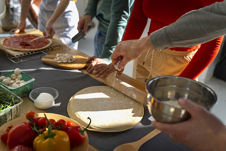 Mehrere Personen bereiten gemeinsam eine Pizza mit frischem Gemüse zu.