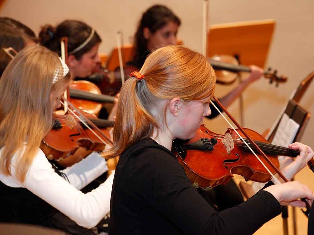 Junge Musikschüler proben auf ihren Violinen, ein Mädchen in Nahaufnahme streicht mit dem Bogen über ihre Geige