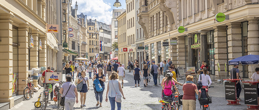 Blick in die Leipziger Straße, links und rechts Einkaufsläden, sommerlich gekleidete Menschen laufen durch die Straße