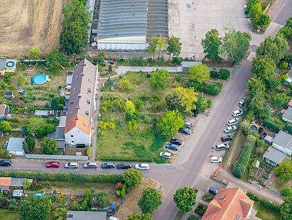 Luftbild einer Grünfläche mit einzelnen Bäumen an einer Straßenkreuzung, links stehen Reihenhäuser, im Hintergrund sieht man Teile einer Lagerhalle