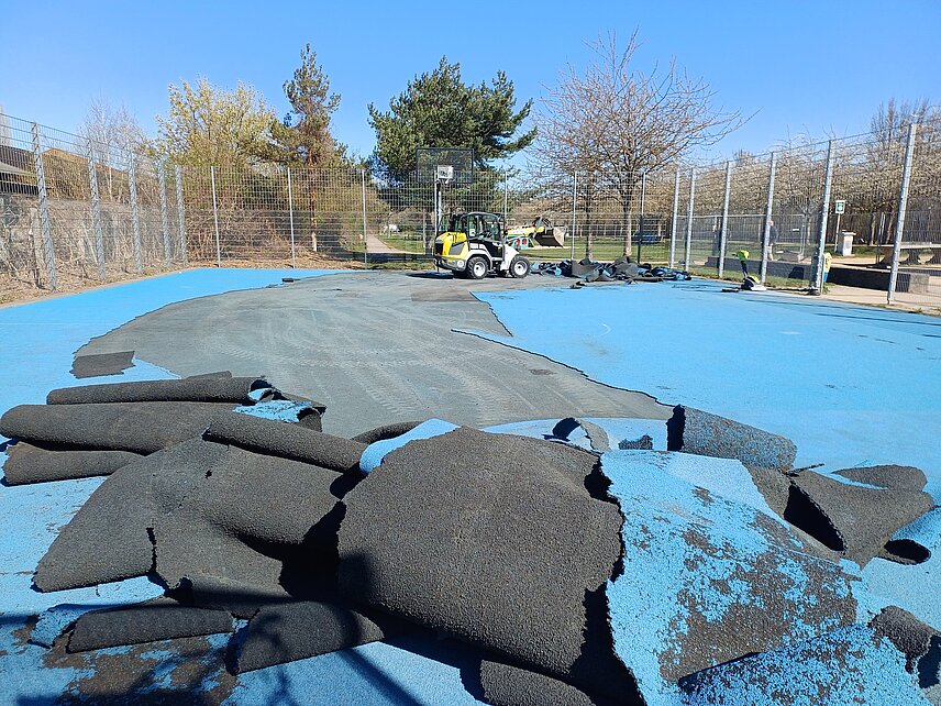Der Belag des Basketballplatzes am Thüringer Bahnhof wird für rund 70.000 Euro erneuert. Im Vordergrund bereits abgerissener Belag, im Hintergrund Zaun und Baumaschine.