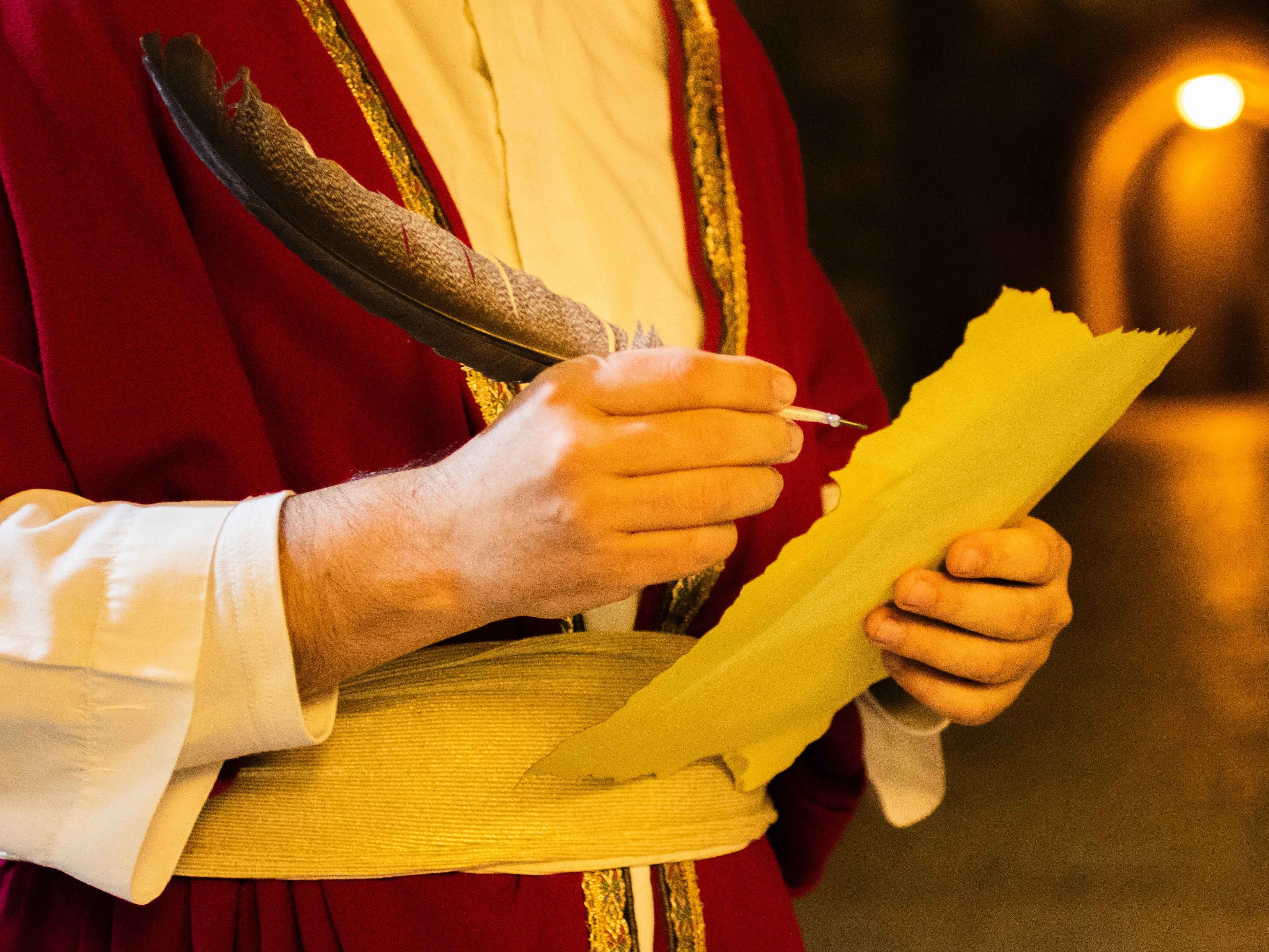 Person in historischer Kleidung hält eine Vogelfeder in der rechten Hand, in der linken Hand hält sie ein Schriftstück