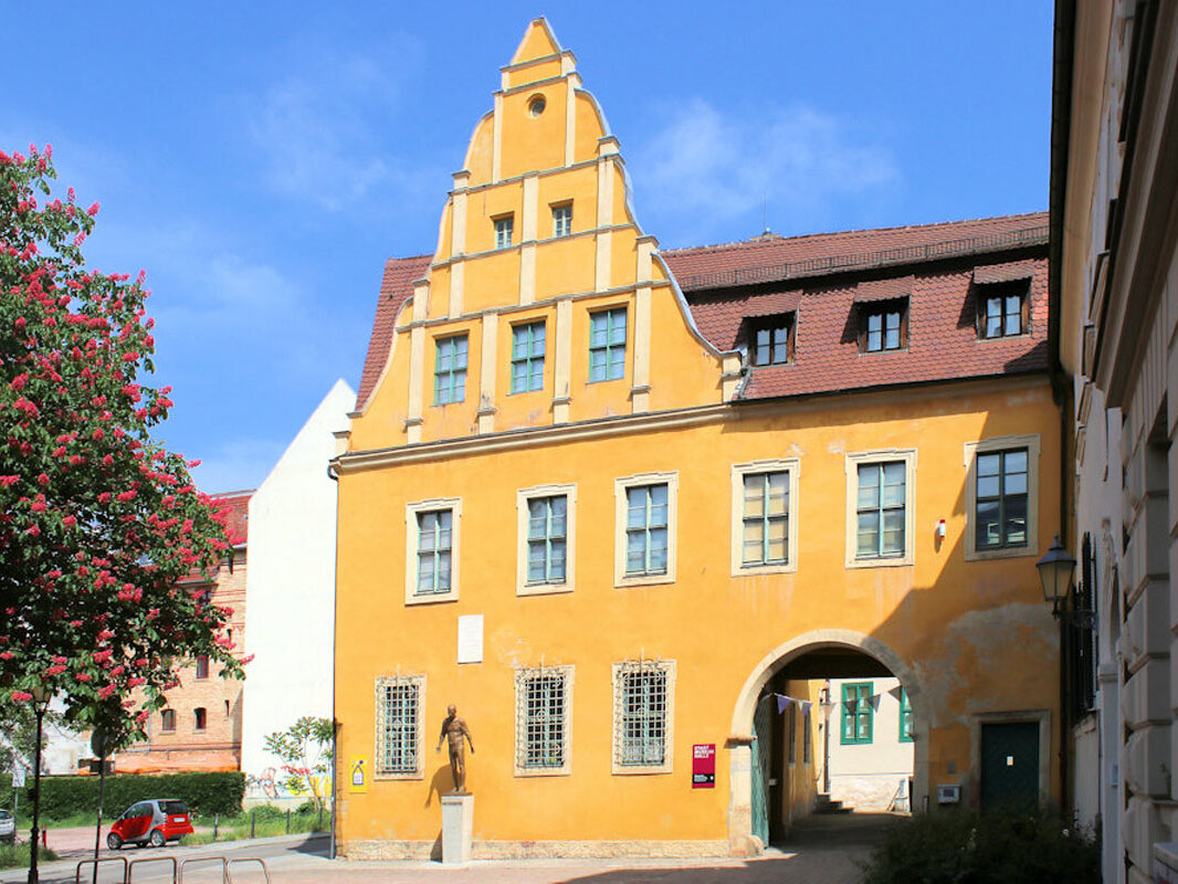 Blick auf einem auffälligen, ockergelb gestalteten Gebäude mit Renaissancegiebeln und großer Toreinfahrt. Im Vordergrund der Platz zum Kleinen Berlin