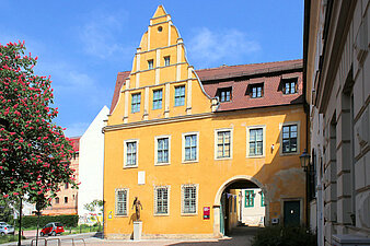 Blick auf einem auffälligen, ockergelb gestalteten Gebäude mit Renaissancegiebeln und großer Toreinfahrt. Im Vordergrund der Platz zum Kleinen Berlin