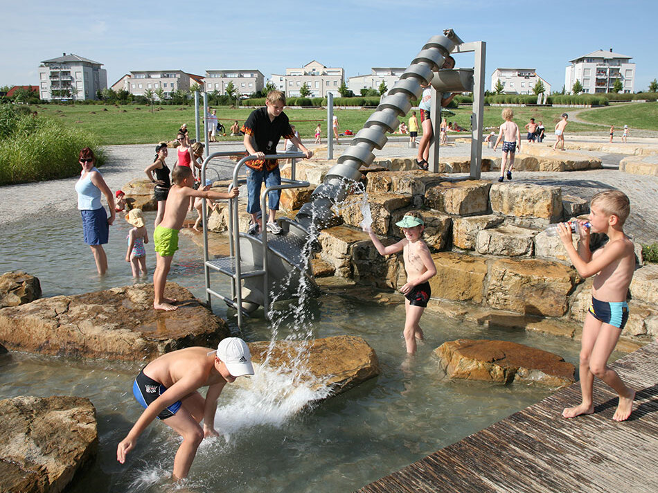 Kinder spielen im Hochsommer auf Wasserspielplatz in Heide-Süd, im Hintergrund Wohnhäuser