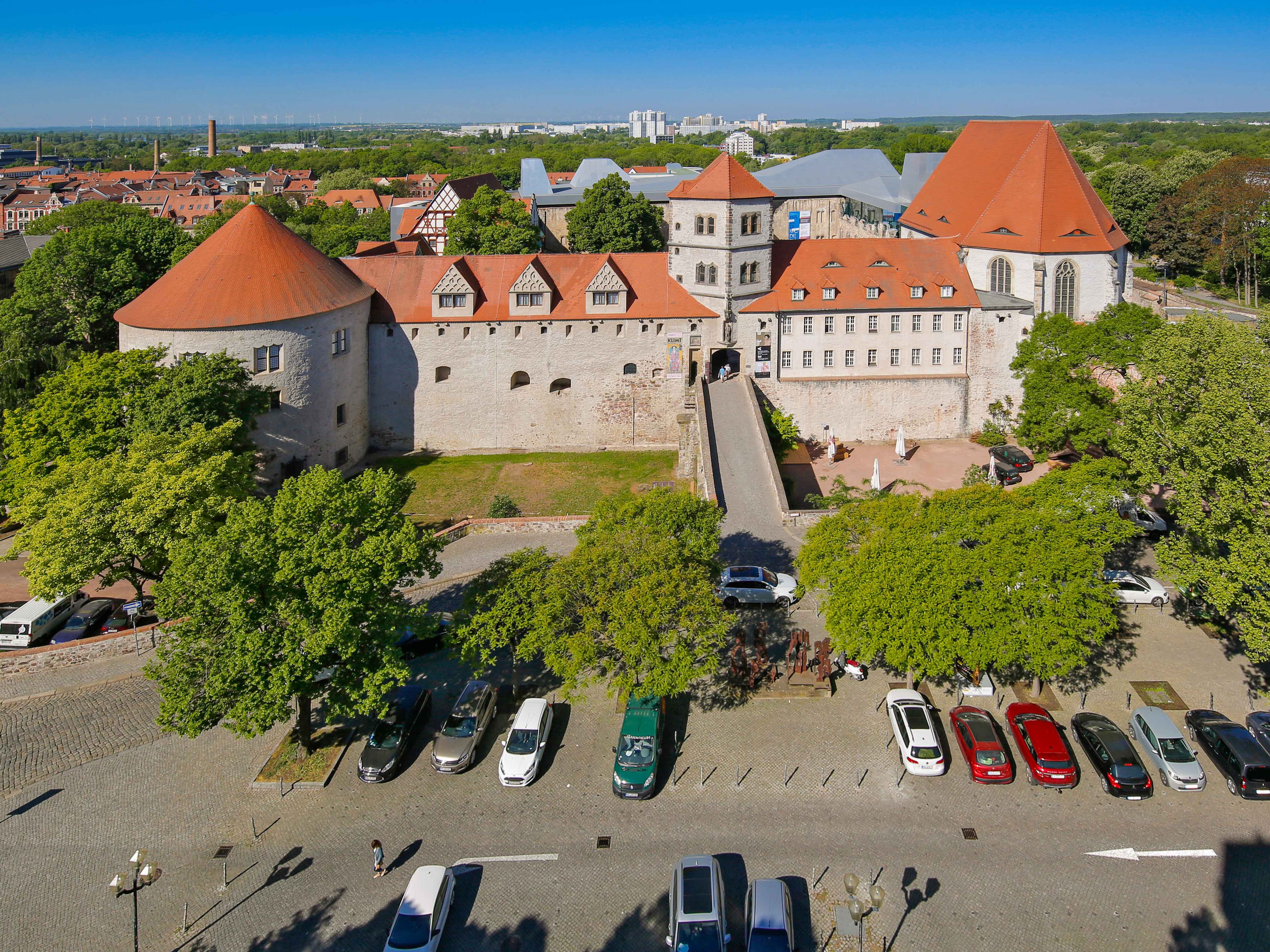 Blick von oben auf die Gemäuer der Moritzburg mit roten Dächern, umrahmt von grün beblätterten Bäumen