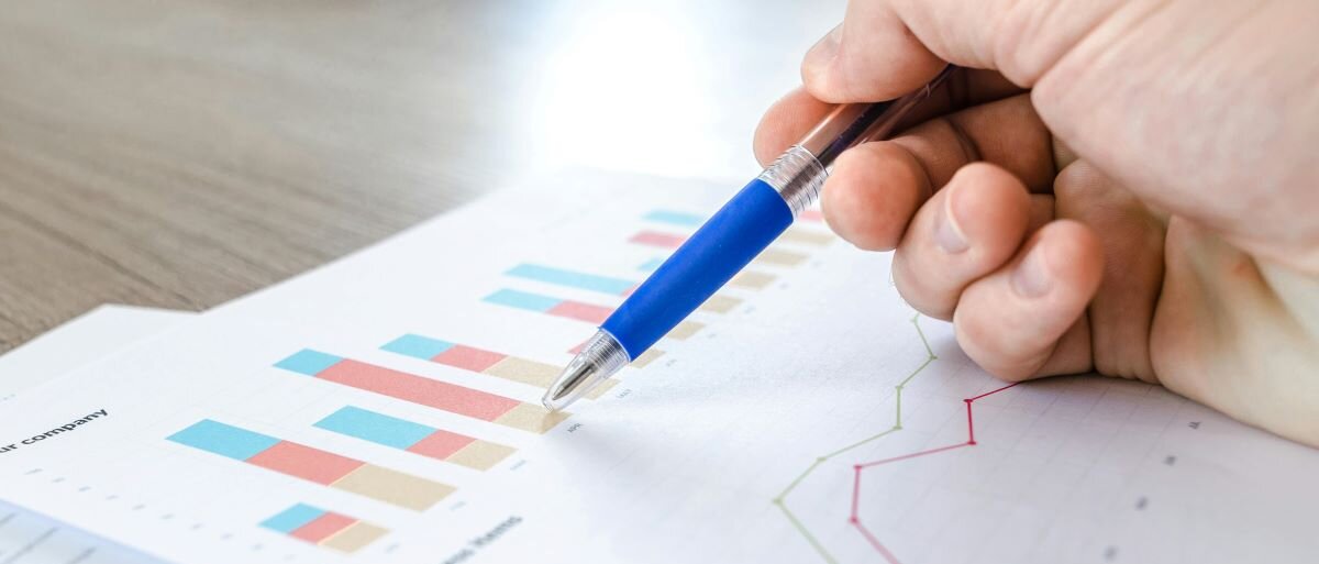 Eine Hand zeigt mit einem blauen Stift auf eines von mehreren Diagrammen, die auf einem Blatt Papier abgebildet sind. Ein weiteres Blatt liegt daneben und enthält Text sowie ein weiteres Diagramm.