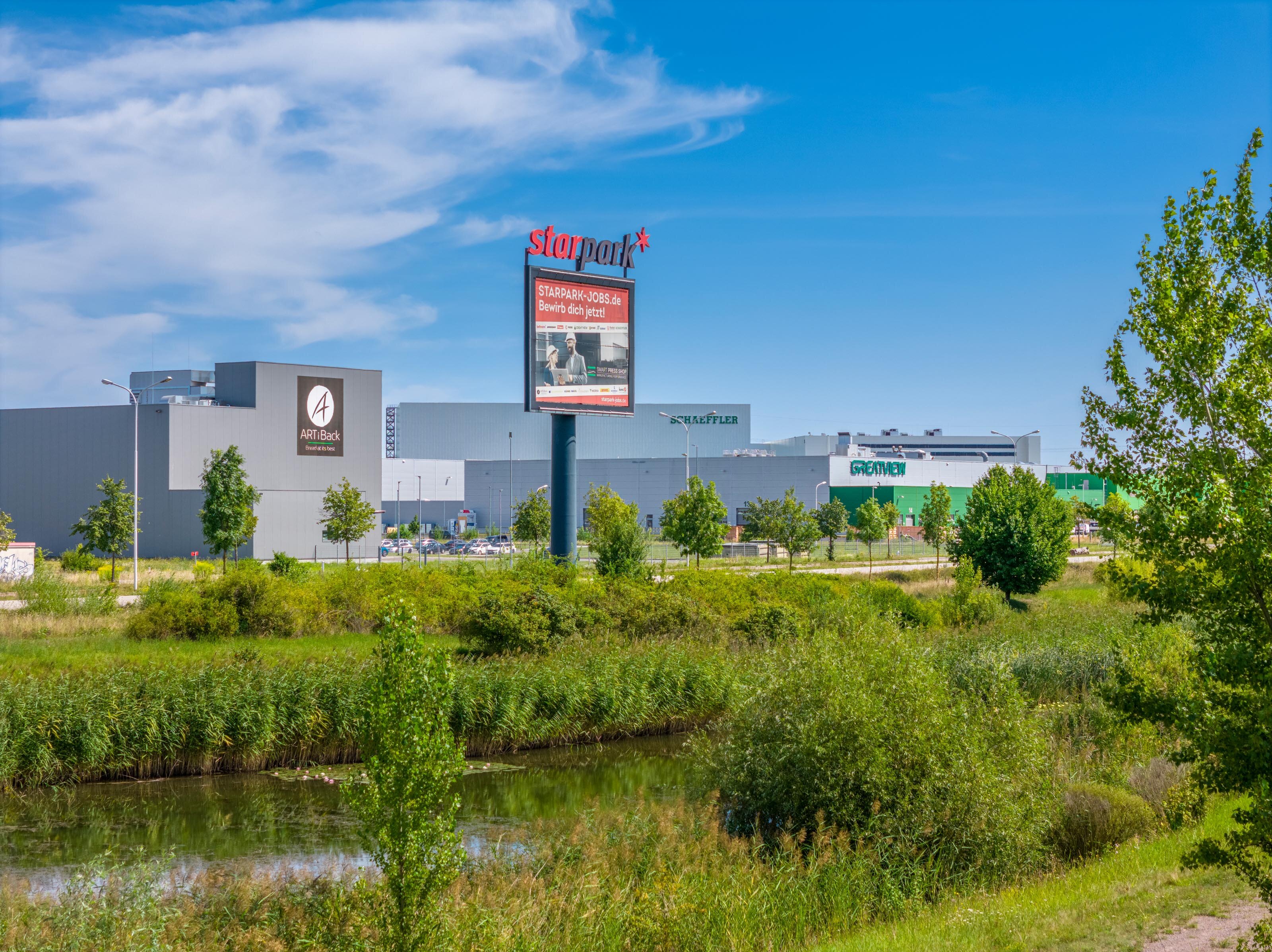 Dieses Bild hat im Vordergrund eine Wiese mit Büschen. Dahinter befindet sich das Werbeschild des hallischen Industrieparks Star Park. Am Horizont können die ersten Unternehmensgebäude des Star Parks gesehen werden.