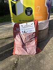 Ein befüllter Müllsack steht an einer Litfaßsäule zur Abholung bereit.