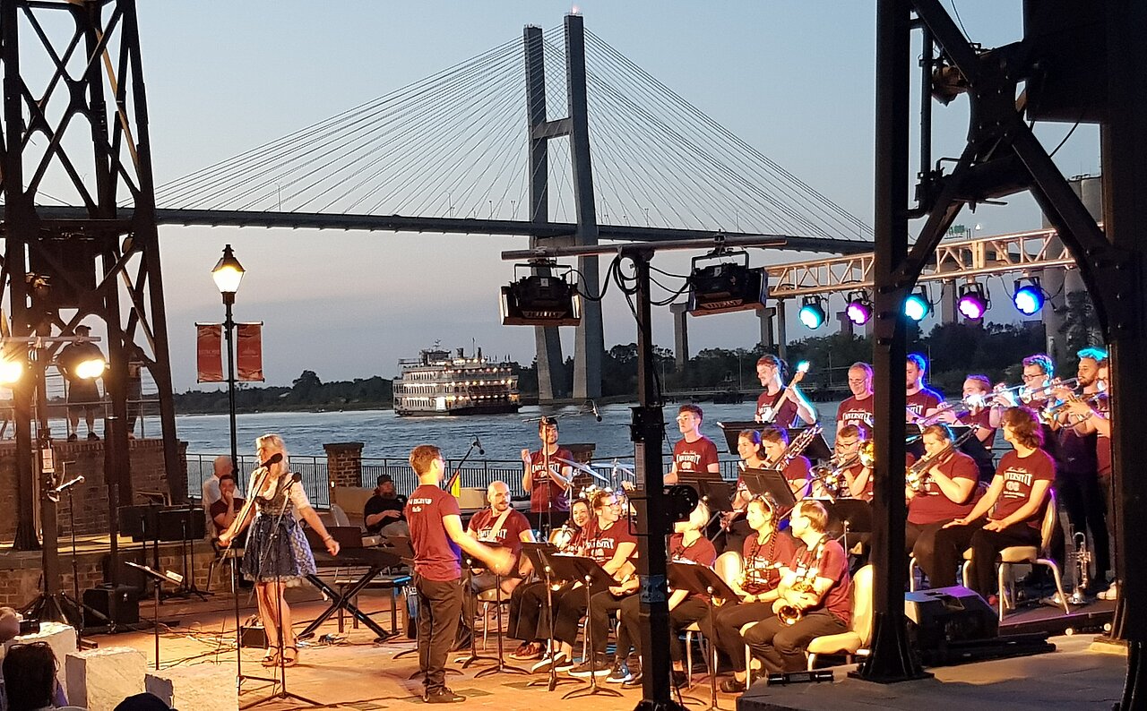 Die Band mit einem Konzert am Fluss. Im Hintergrund eine große Brücke und das Schiff Georgia Queen