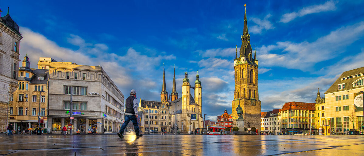 Marktplatz mit Blick auf die fünf Türme. Im Vordergrund nasses Pflaster, ein Passant läuft Richtung Händel-Denkmal