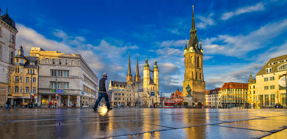 Marktplatz mit Blick auf die fünf Türme. Im Vordergrund nasses Pflaster, ein Passant läuft Richtung Händel-Denkmal