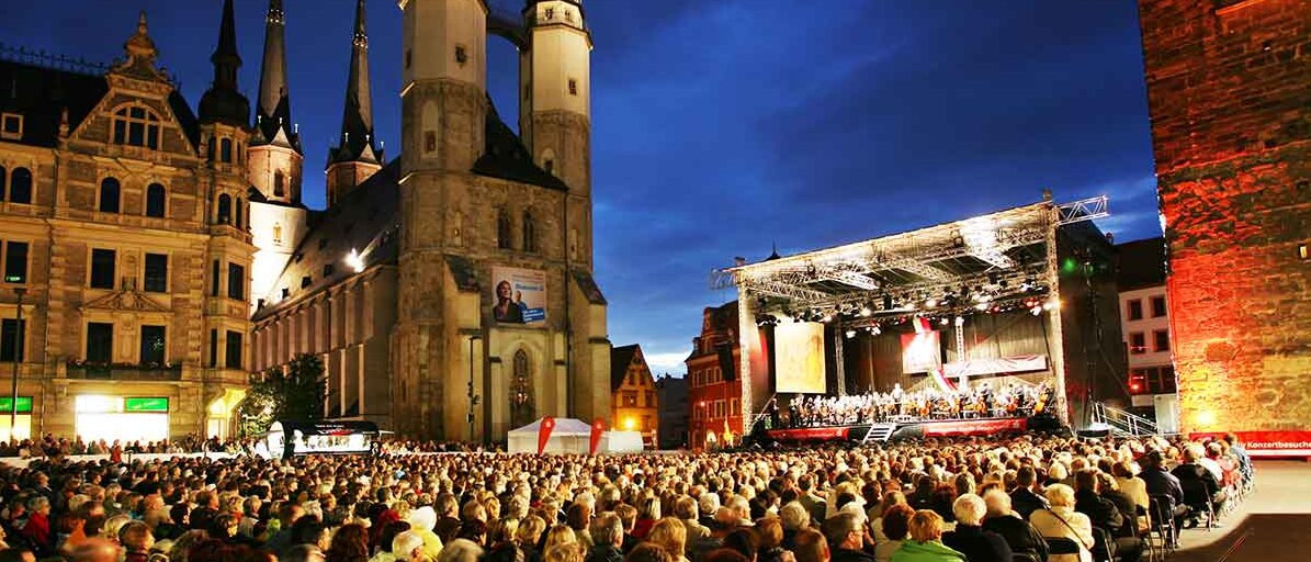 Sitzende Zuschauer vor einer Bühne, aufgestellt neben dem Roten Turm auf dem Marktplatz bei abendlicher Stimmung