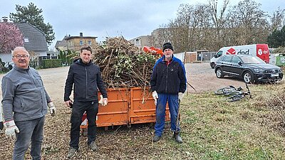 Drei Beteiligte stehen vor einem mit Gehölzen befüllten Container.