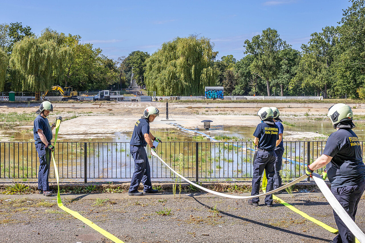 Feuerwehrmänner stehen mit Schläuchen in der Hand am Ufer des noch leeren Fontäne-Teich