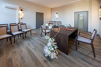 Blick in das Eheschließungszimmer im Ratshof, 13 leere Stühle, vorn ein Herz mit weißen Blumen, brauner Schreibtisch, Laminatfußboden