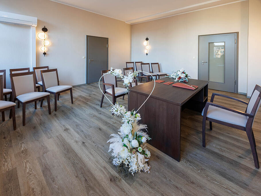 Blick in das Eheschließungszimmer im Ratshof, 13 leere Stühle, vorn ein Herz mit weißen Blumen, brauner Schreibtisch, Laminatfußboden