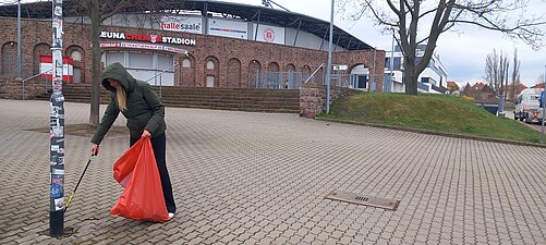 Eine Frau sammelt Streumüll mit einer Greifzange auf. Im Hintergrund ist das Stadion zu erkennen.