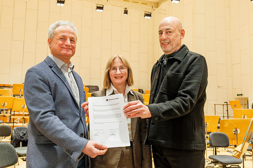 Dirigent Fabrice Bollon (links) und TOOH-Geschäftsführer Klaus Dörr halten Vertrag.  Hinter ihnen steht die Beigeordnete für Kultur und Sport, Dr. Judith Marquardt.