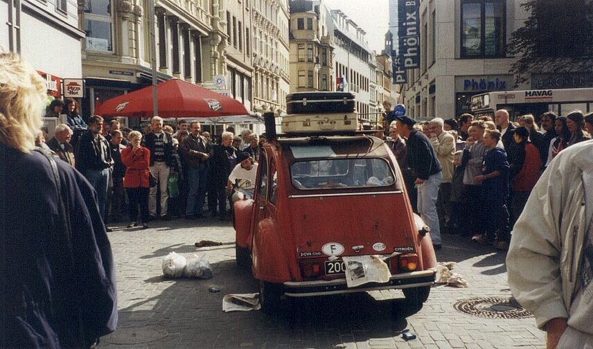 Straßentheater - altes Auto (Ente) steht in der Fußgängerzone von Passanten umringt 