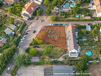 Luftbildaufnahme einer Einfamilienhaussiedlung - rechts Reihenhausbebauung mit Gärten, auf der linken Seite getrennt durch eine Straße Kleingärten, die auf einer Grünfläche liegende Verkaufsparzelle ist farbig markiert
