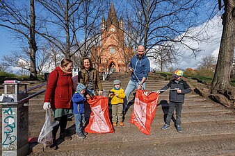 Im Vordergrund stehen mehrere befüllte Müllsäcke und im Hintergrund sind Kinder und Erwachsene zu sehen.