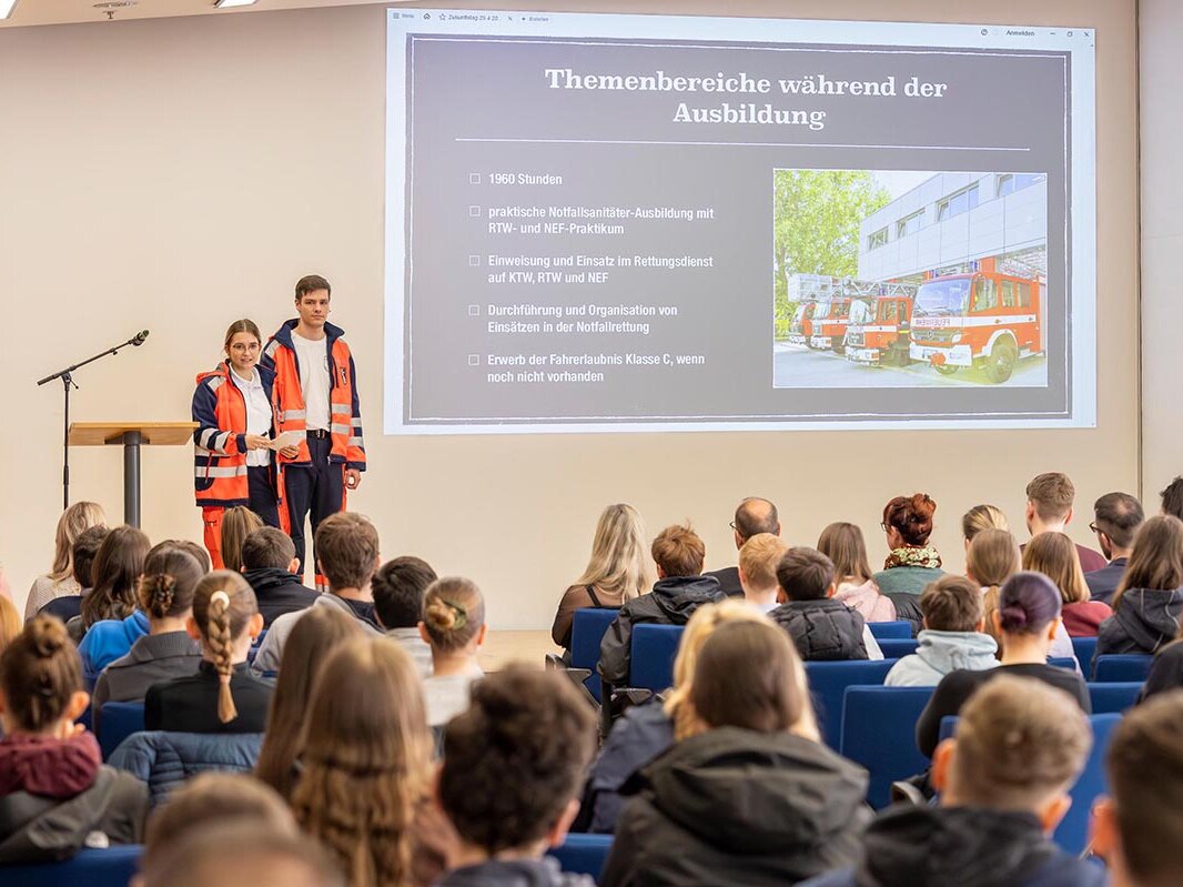 2 Azubis (1 Mädchen und 1 Junge) erklären auf einer Bühne mit Beamer die Themenbereiche während der Ausbildung zum Notfallsanitäter