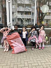 Fünf freudige Kinder stehen vor der Kita und geben Handzeichen. Ein befüllter Müllsack wird von einem Kind festgehalten.