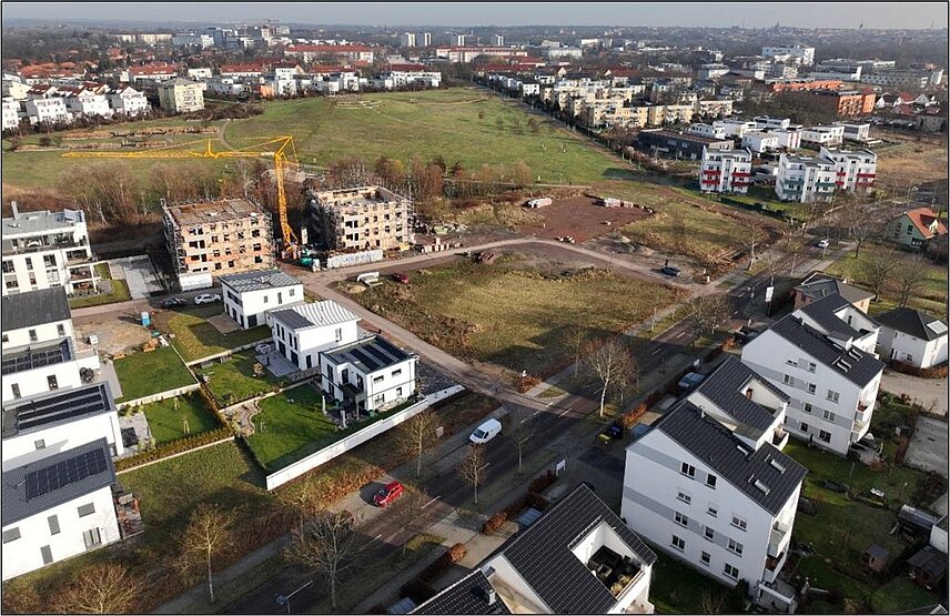 Blick über einen Teil des Baugebietes Heide-Süd, vorn rechts mehrgeschossige moderne Mehrfamlilienhäusern, links neie Einfamilienhäuser, dazwischen eine Straße mit Bäumen, mittig sind freie Bauflächen zu sehen, links davon zwei Wohngebäude, die noch noch im Bau sind, dazwischen steht ein großer Kran, daran anschließend grüne Wiesen, das sogenannte Grüne Dreieck, im Hintergrund stehen viele neue Mehrfamilienhäuser mit unterschiedlichen Fassaden, am Horizont kann man die Altstadt von Halle erkennen