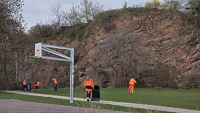 Ein Basketballfeld und im Hintergrund sind Personen mit Warnwesten, die Müll aufsammeln.