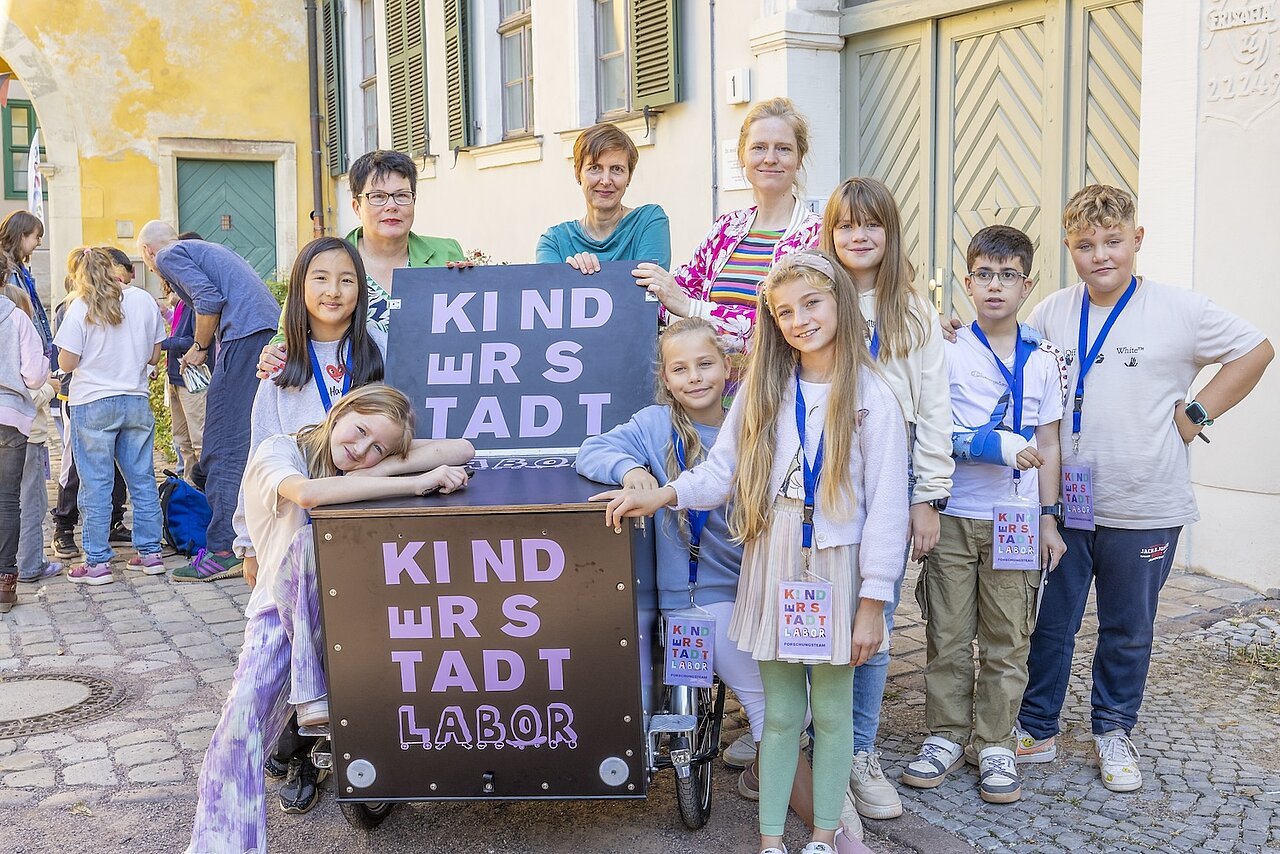 Die drei Personen stehen mit einer Kindergruppe vor zwei Schildern für das Labor.