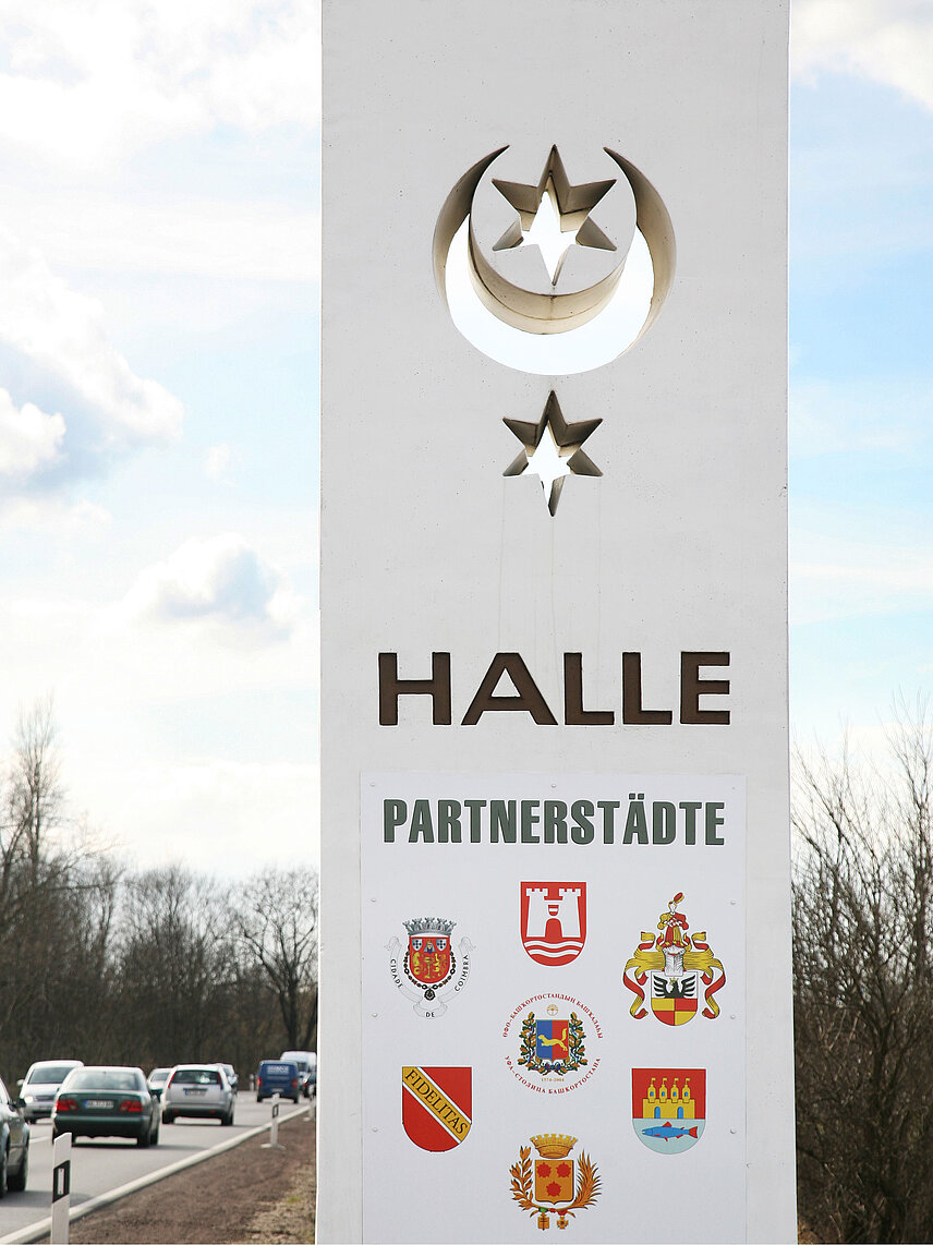 Weiße Stehle mit Wappen der Stadt Halle, Schriftzug Halle Partnerstädte und bunten Wappen der Partnerstädte, steht am Ortseingang der Bundesstraße 6