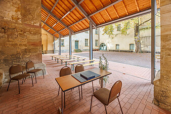 Bild mit Tischen, Stühlen und Bänken und einer Schmuckvase auf rotem Steinboden unter einer an die Außenwand der Ulrichskirche angebrachte Überdachung, mit Blick in den Innenhof der Ulrichskirche. Im Hintergrund eine Kastanie.
