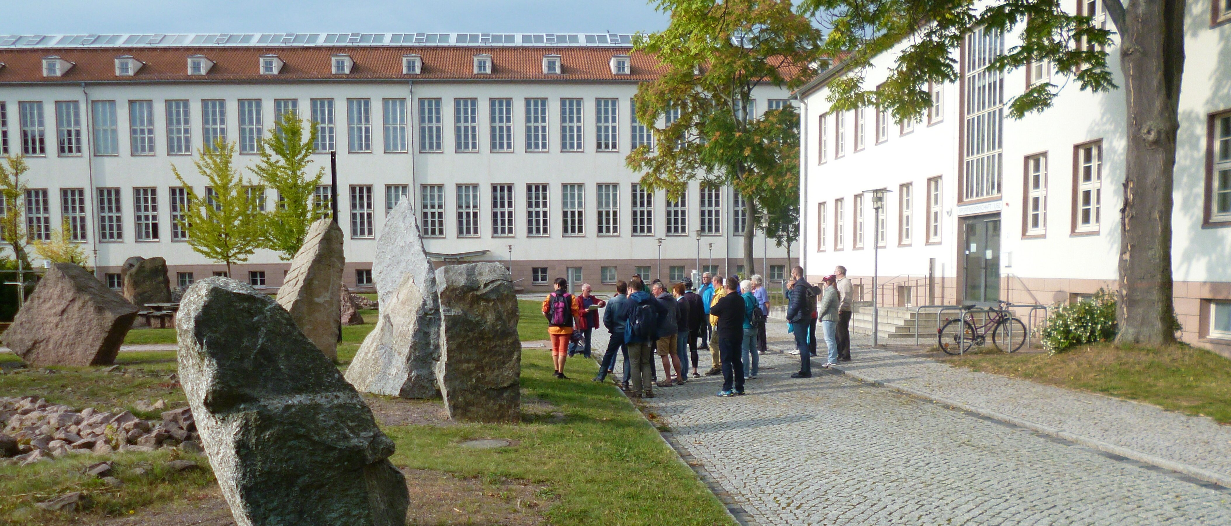 Menschengruppe vor großen Steinen verschiedener Gesteinsarten, im Hintergrund Universitätsgebäude
