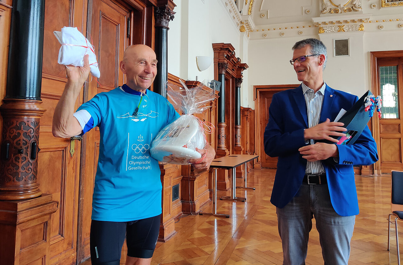 Bürgermeister Egbert Geier begrüßt den Initiator des „Rollenden Klassenzimmers“, Ortwin Czarnowski, im Stadthaus. Beide halten Gastgeschenke in der Hand.