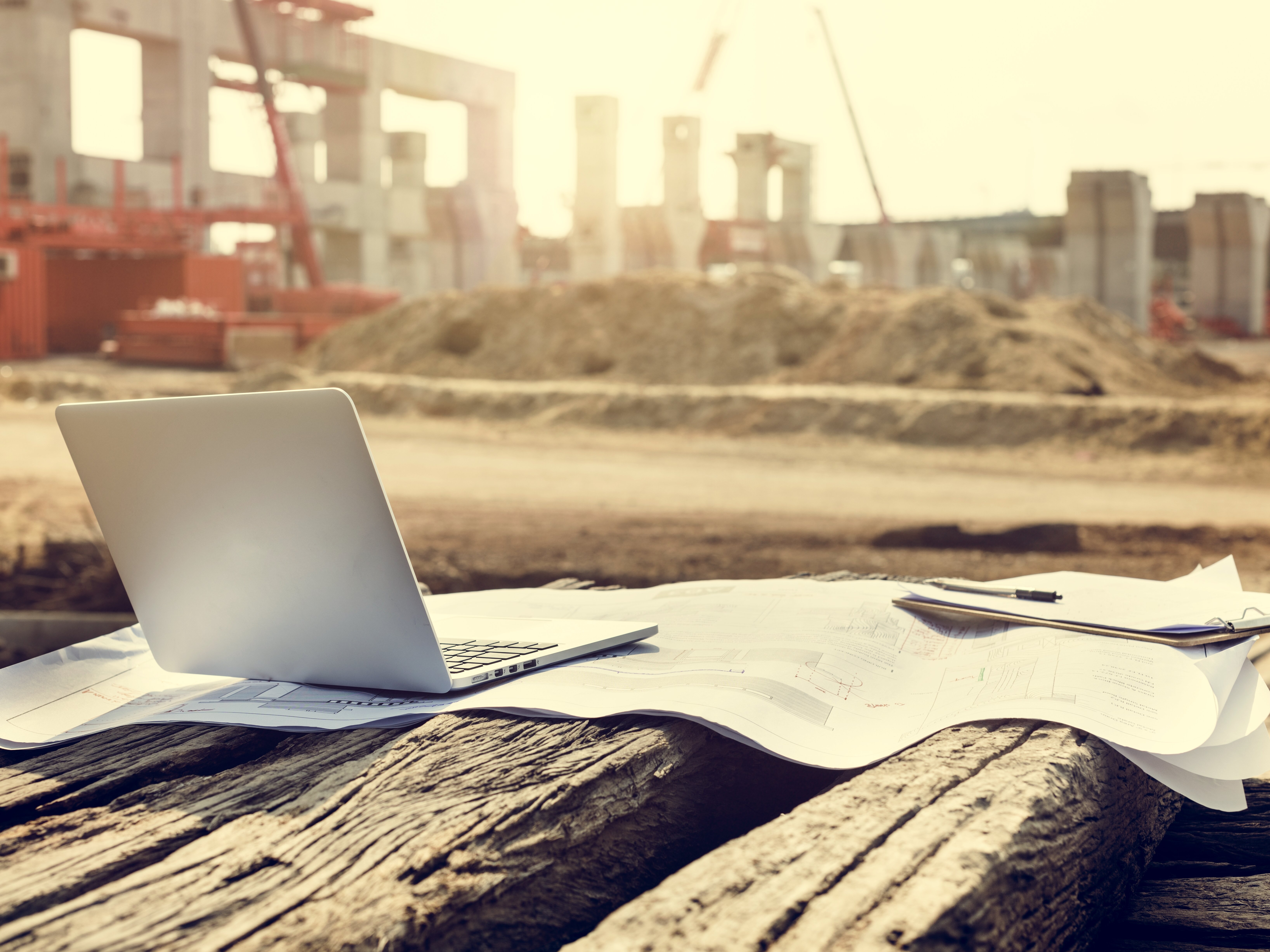 Ein Laptop steht auf einem Bauplan, im Hintergrund ist eine Baustelle zu sehen.